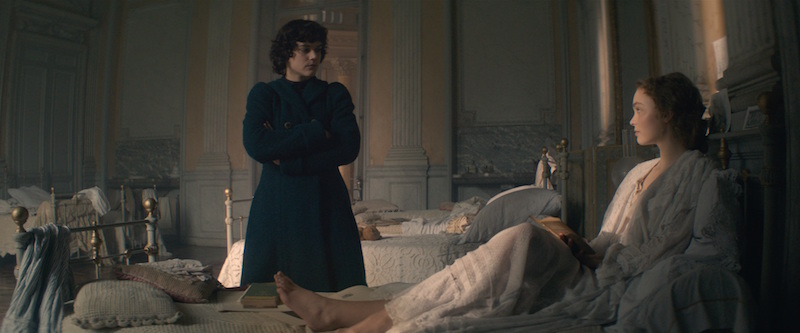 A still of an opulent French bedroom. A person in a dark overcoat looks down at a woman in bed wearing a dressing gown.